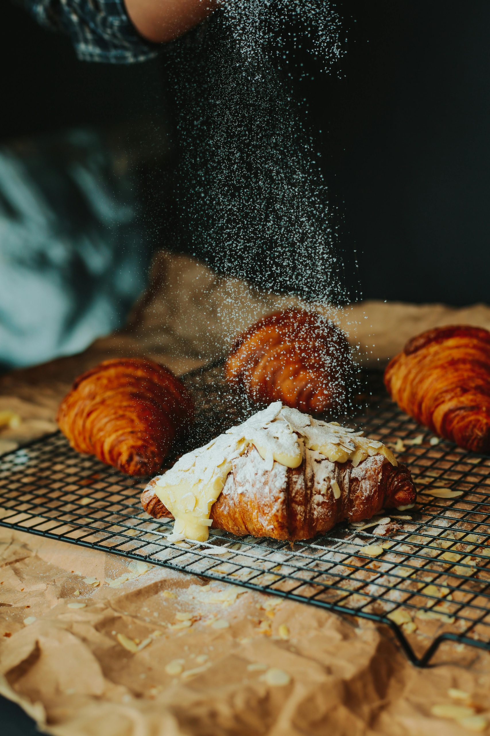 Freshly baked croissants being dusted with powdered sugar on a cooling rack.